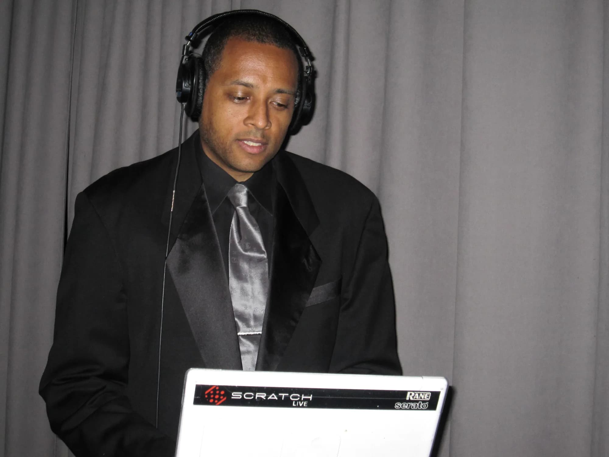 DJ David Raimer cueing a track on headphones over a Serato Scratch Live setup in a black tuxedo during a formal celebration