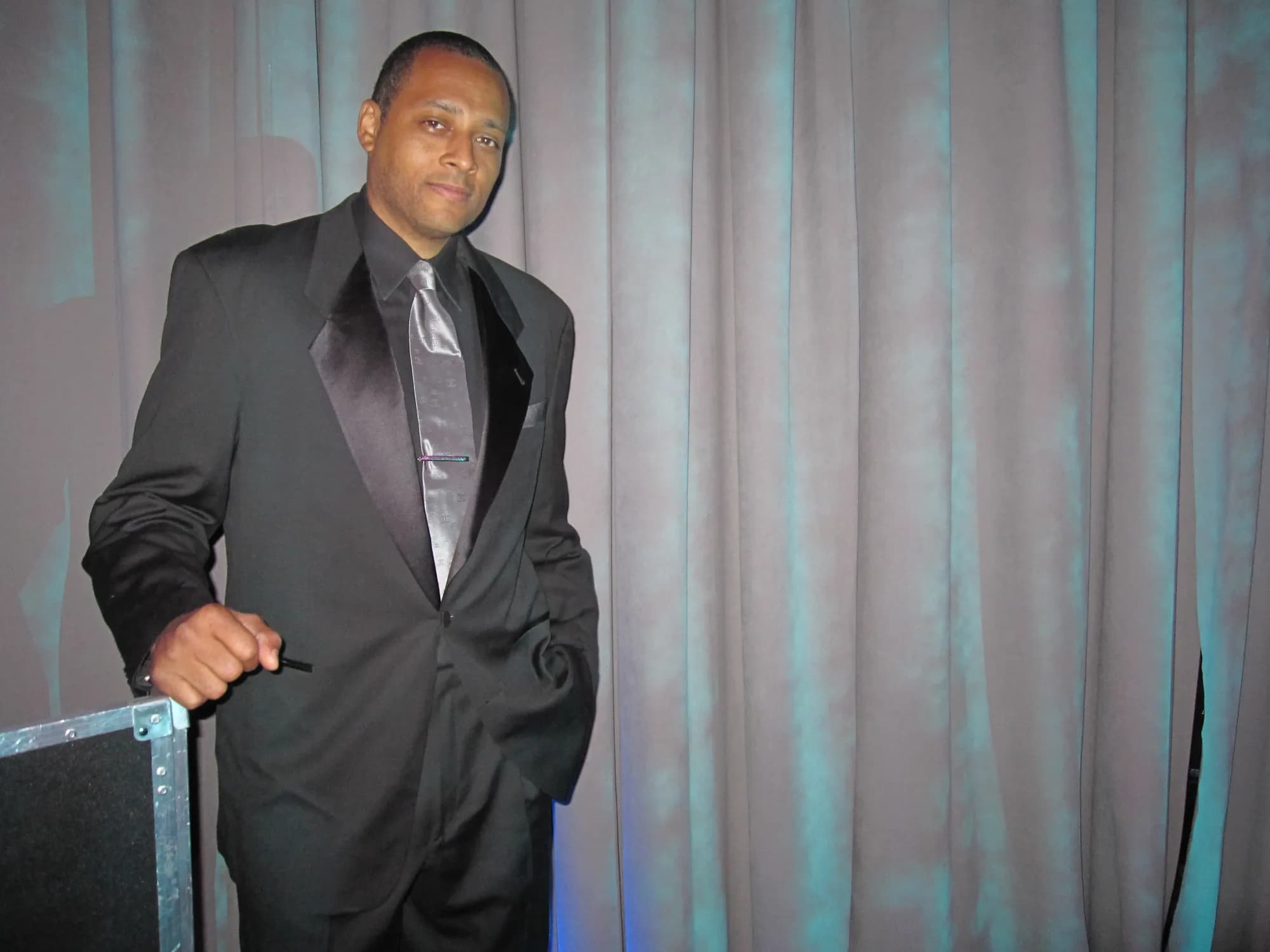 DJ David Raimer in a black tuxedo and silver tie beside his road case at an upscale NYC evening event