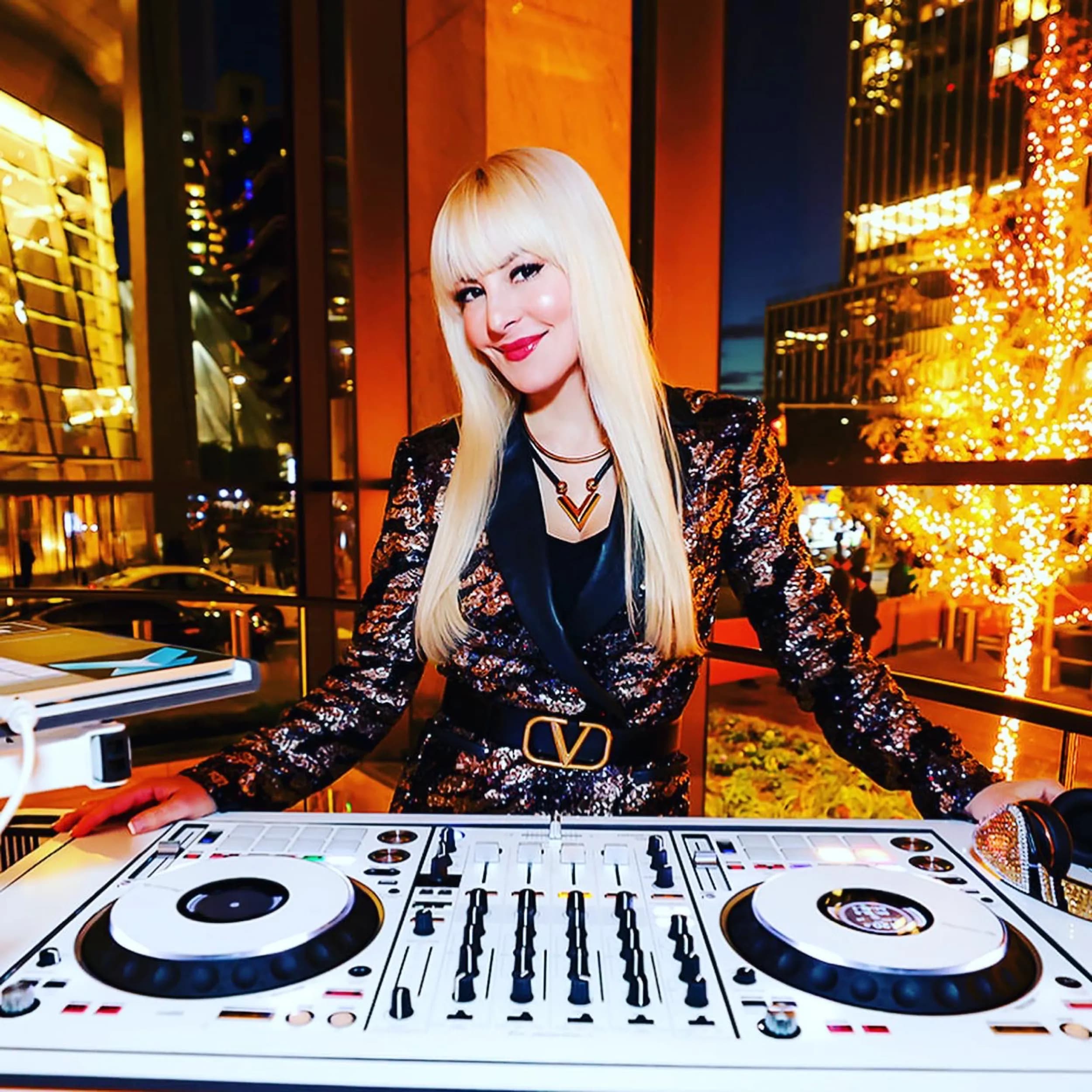DJ Kristaval at the decks against the lit NYC skyline and a holiday tree at a rooftop celebration
