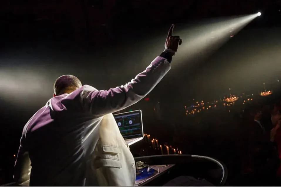 DJ Neza with his hand raised under a single spotlight before a large candlelit crowd at an arena-scale event