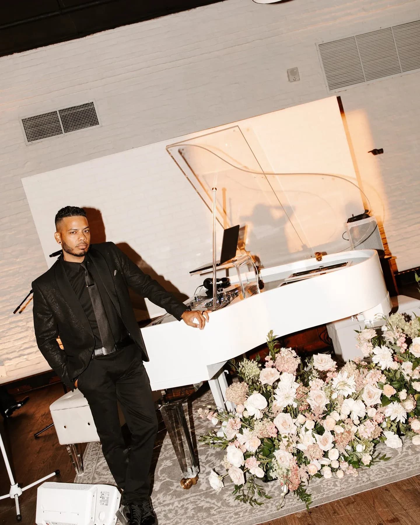 DJ Neza in a black suit beside his signature white baby grand piano DJ booth with cascading florals at an upscale New York wedding