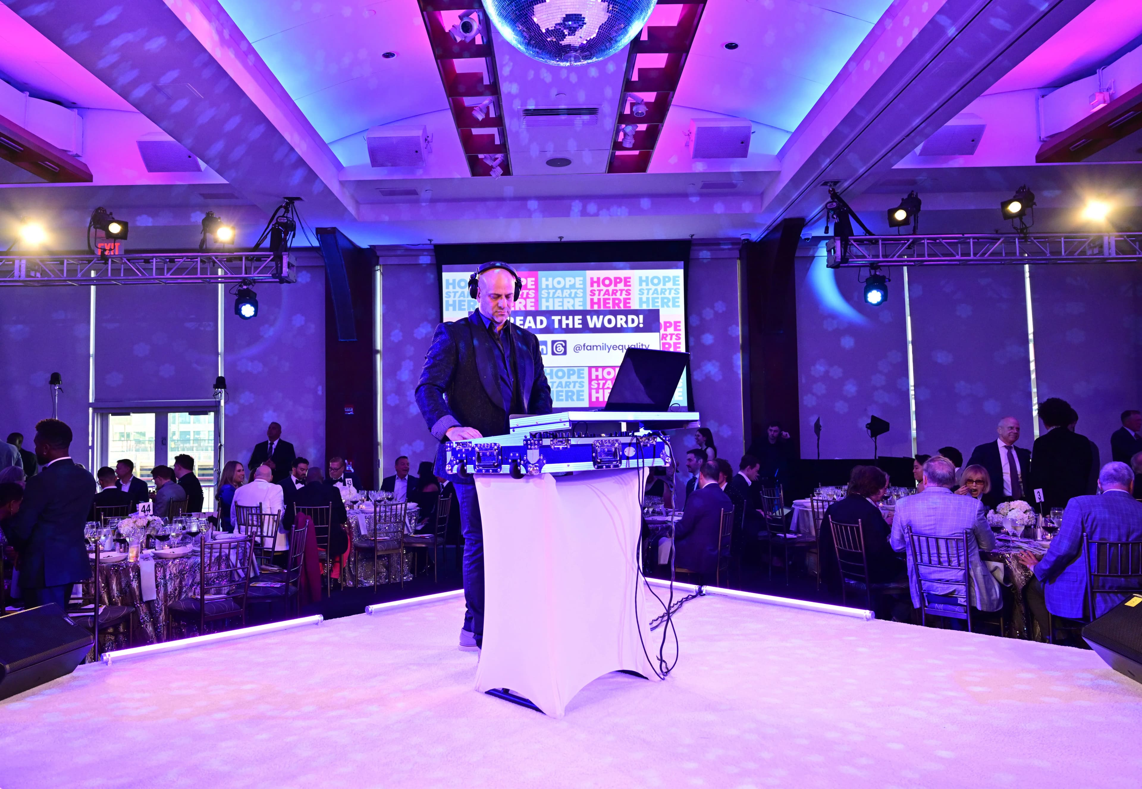 Black-tie charity gala ballroom with dressed guests at banquet tables, uplighting, and produced stage