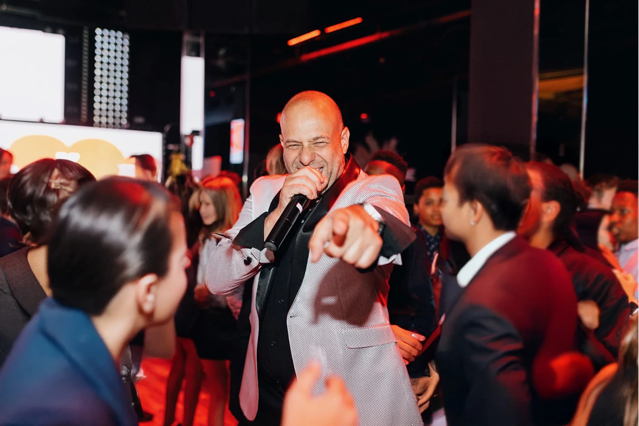 MC Shiran Nicholson laughing on the mic surrounded by a crowd at an upscale event under warm red light