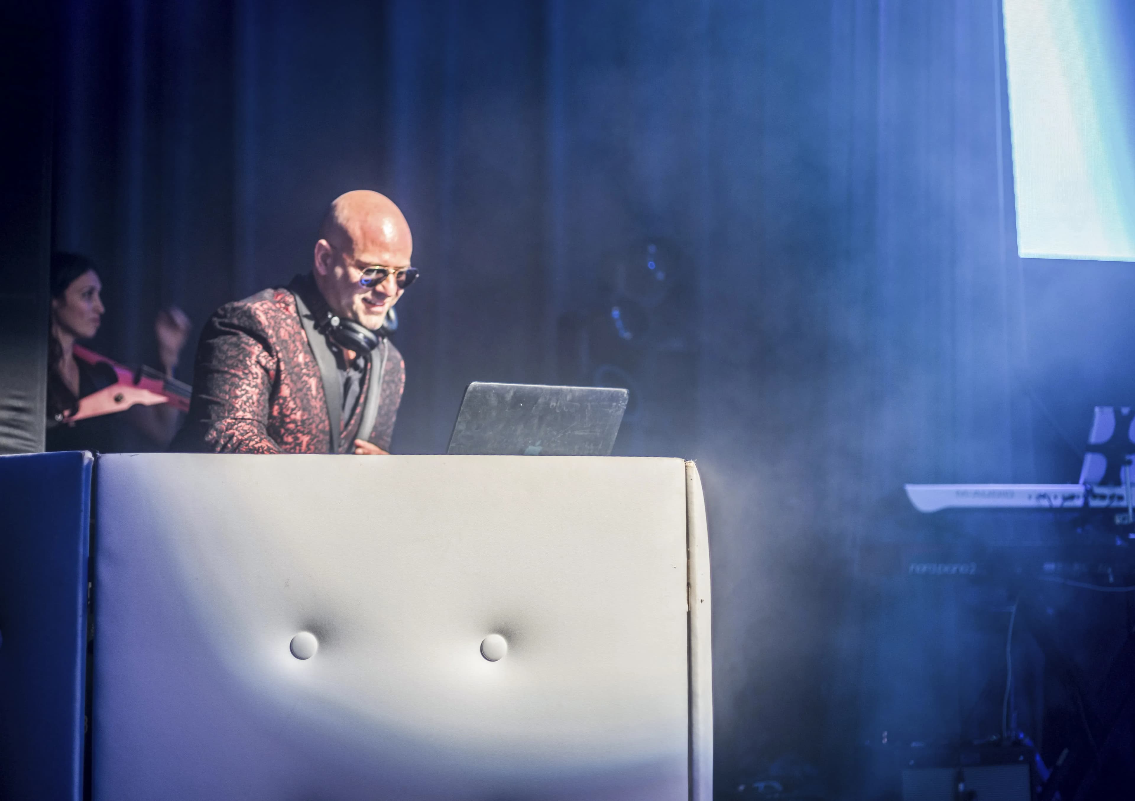 Shiran Nicholson at the DJ booth in a red and black patterned blazer under moody blue haze at a production event