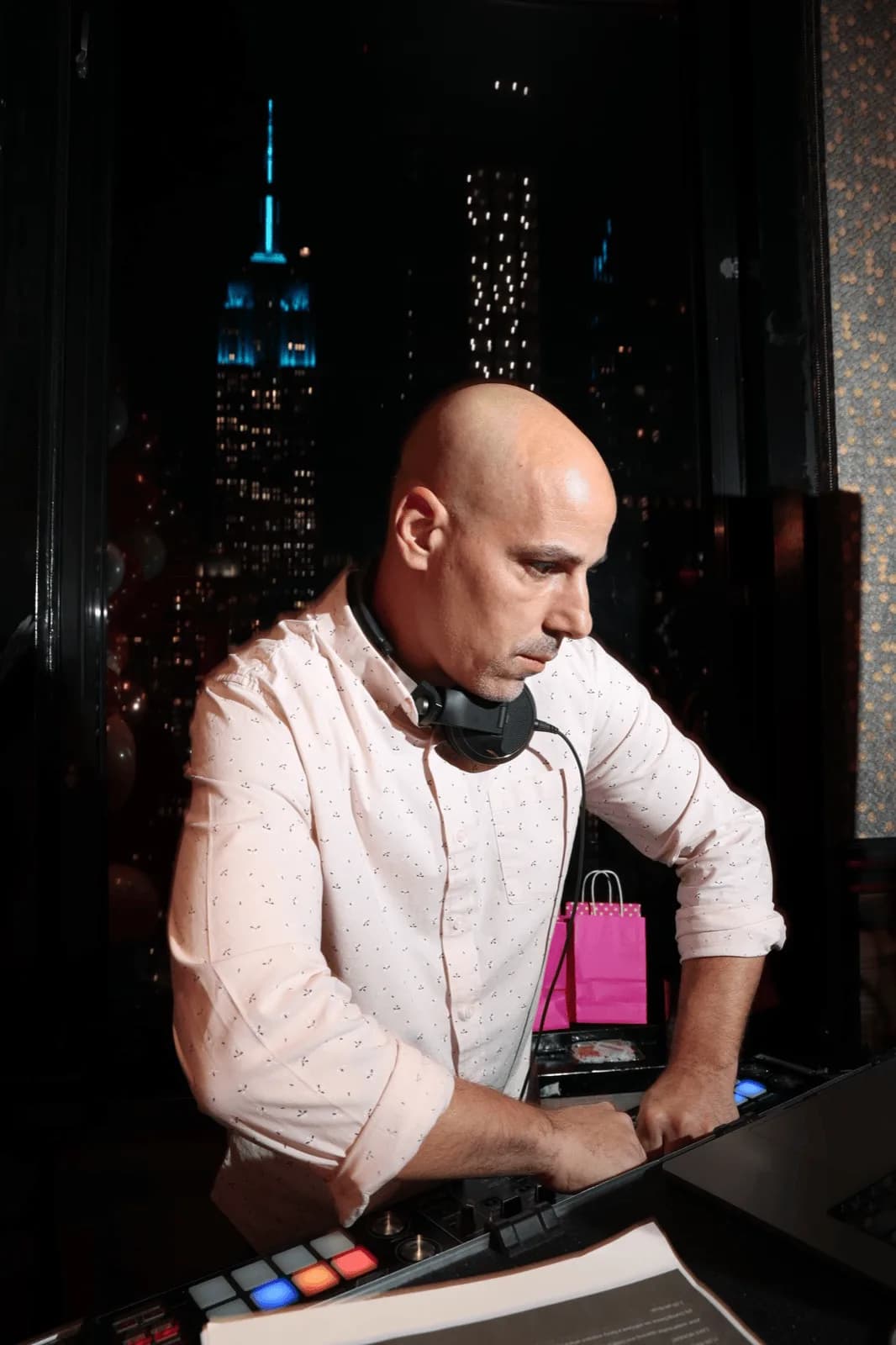 DJ Tommy with arm extended over the decks at a NYC event, the Empire State Building visible through the window behind him