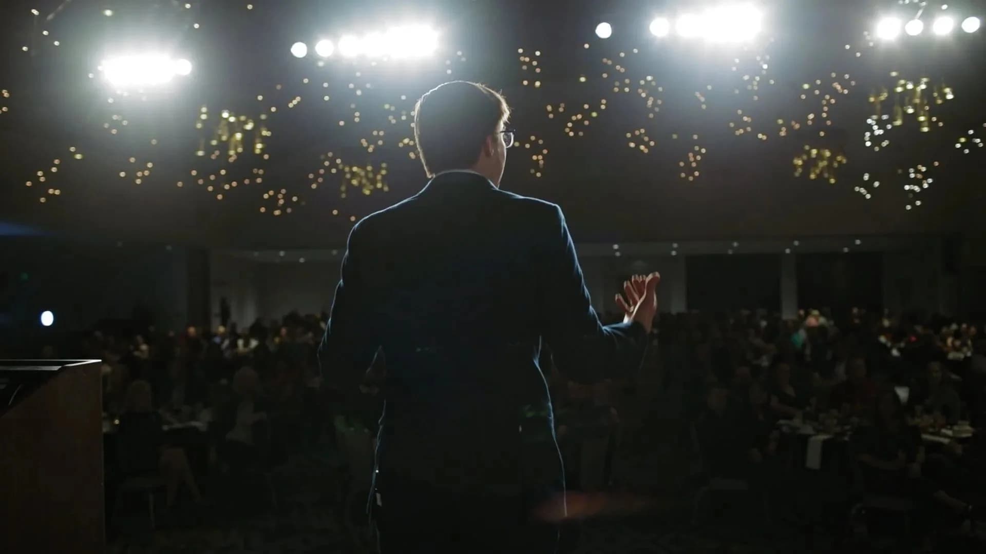 Magician performing on stage to a packed gala audience under warm string lights
