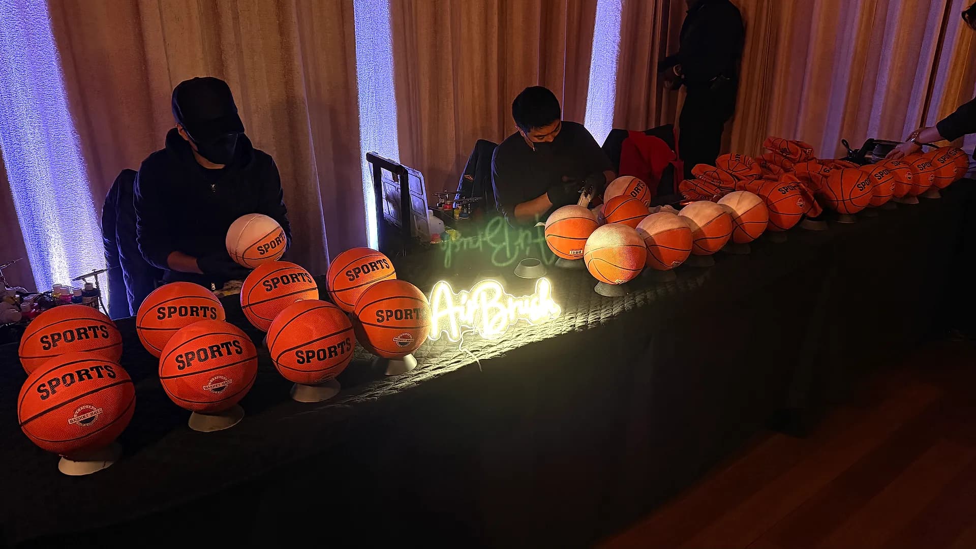 Live airbrush artists customizing sports basketballs at an event station with a glowing AirBrush neon sign
