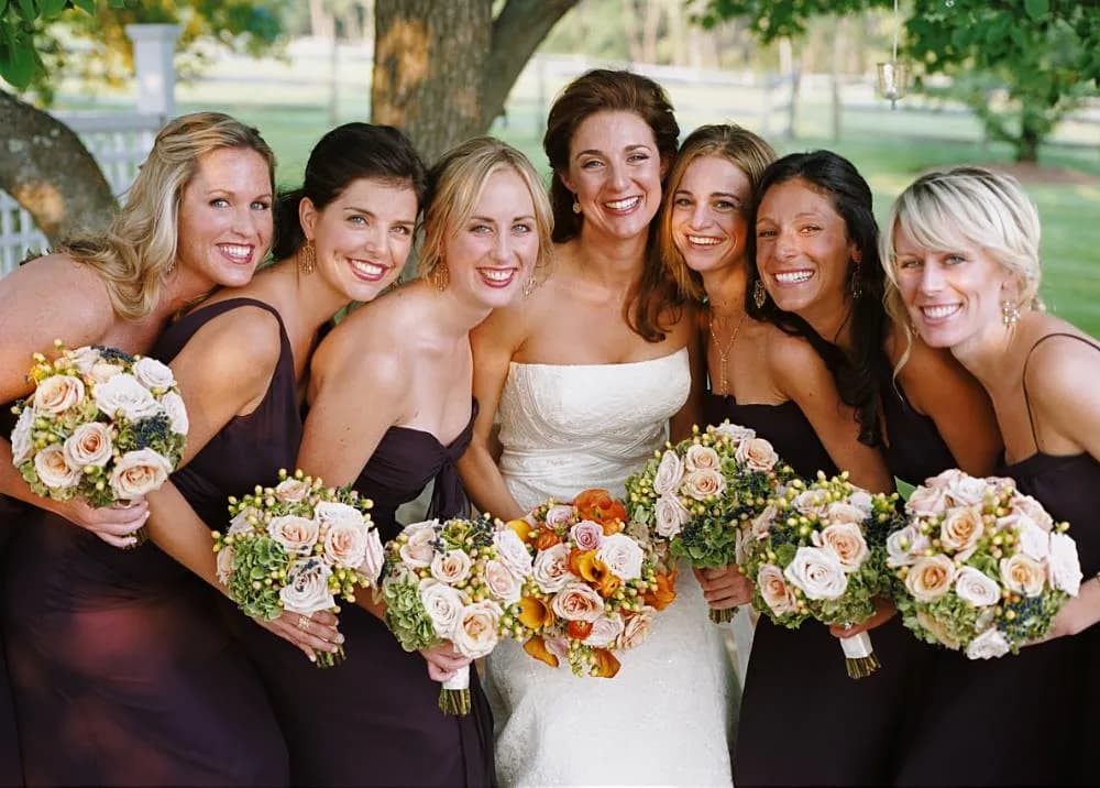 Bride with her bridesmaids in plum dresses holding blush rose bouquets under a tree