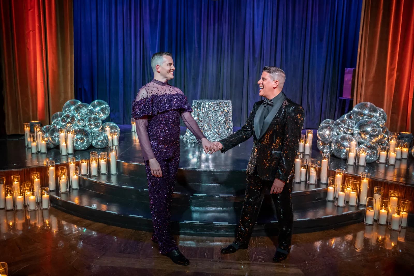 Two grooms in sequined suits holding hands on a candlelit stage surrounded by disco balls